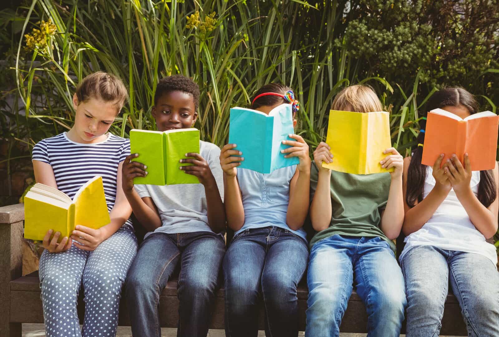 Five children sitting in a row on an outdoor bench, each reading a brightly coloured book